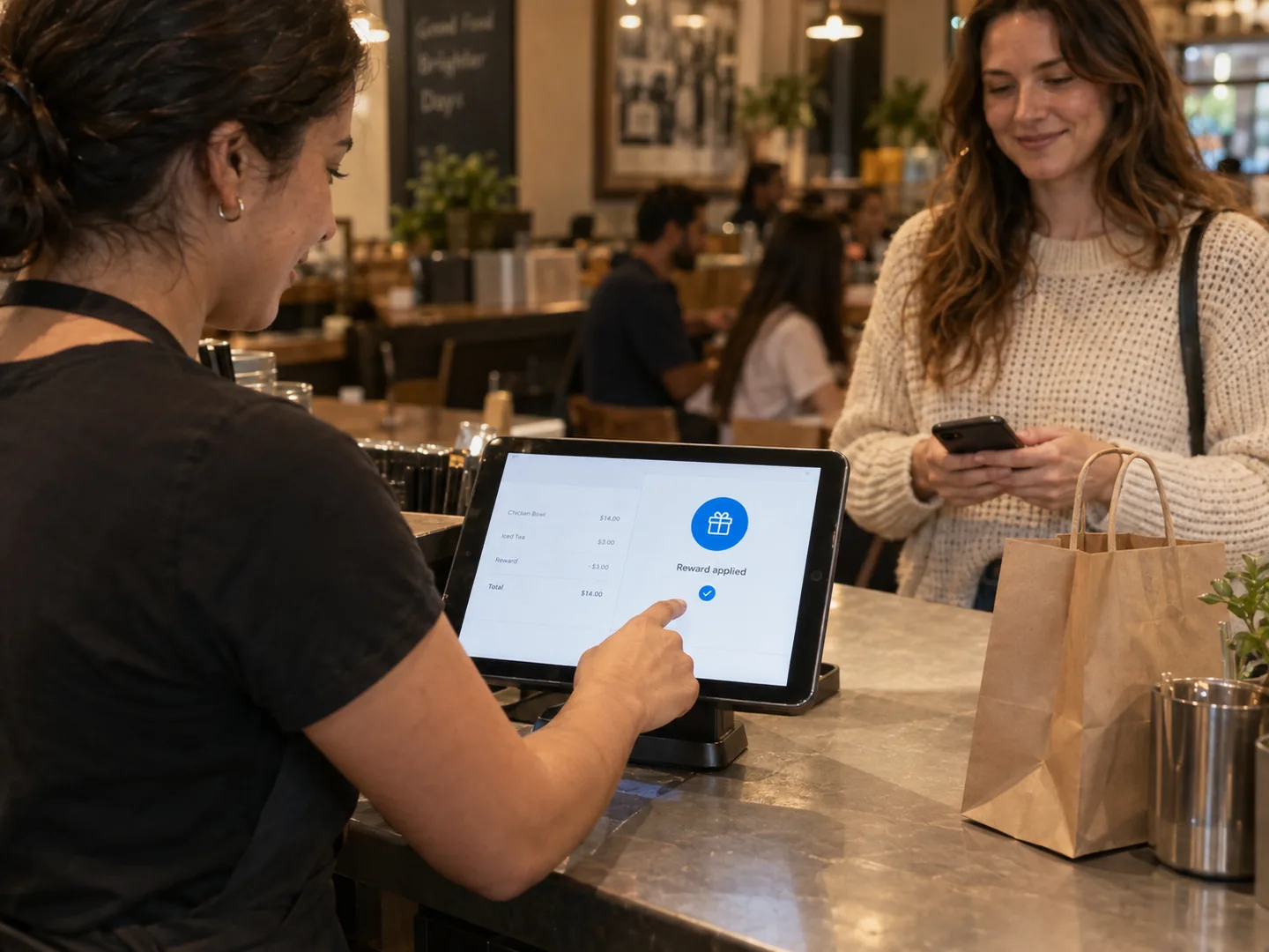 A cashier applying a regular’s reward on the counter tablet while the guest waits with her phone and her order